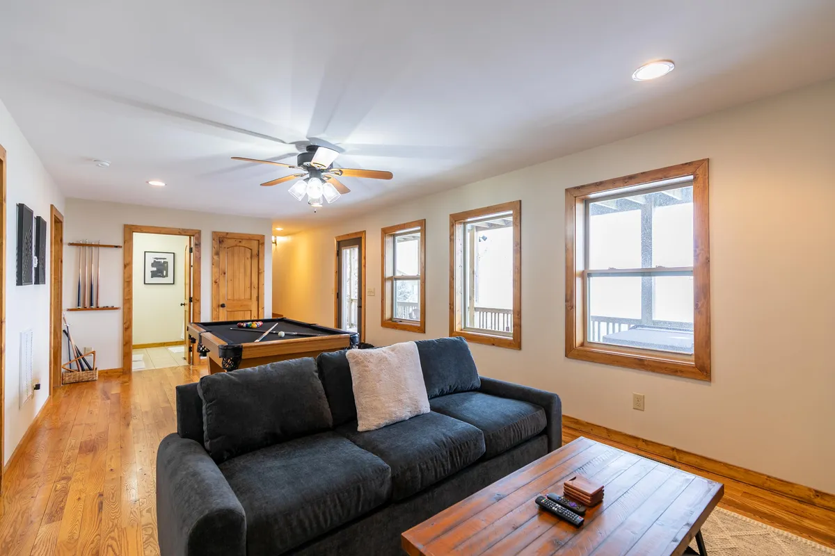 An inviting, casual game room with a pool table, cozy sofa, wooden coffee table, and windows letting in natural light. The atmosphere is relaxed and perfect for both lively games and quiet downtime.