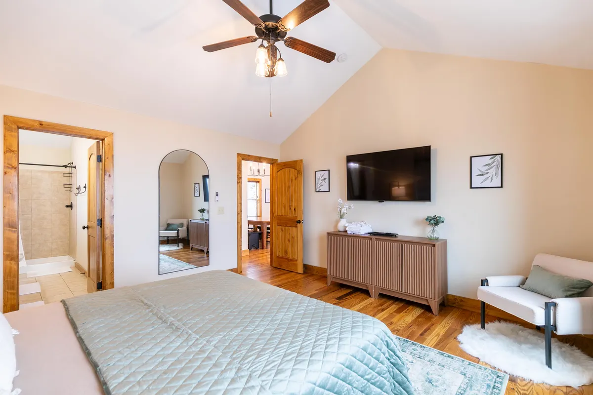 A stylish King bedroom with a comfortable bed, a mounted TV, a sunlit window, a wall mirror, and an inviting armchair. The room feels cozy and well-appointed, suggesting modern comforts and space for both relaxation and entertainment.