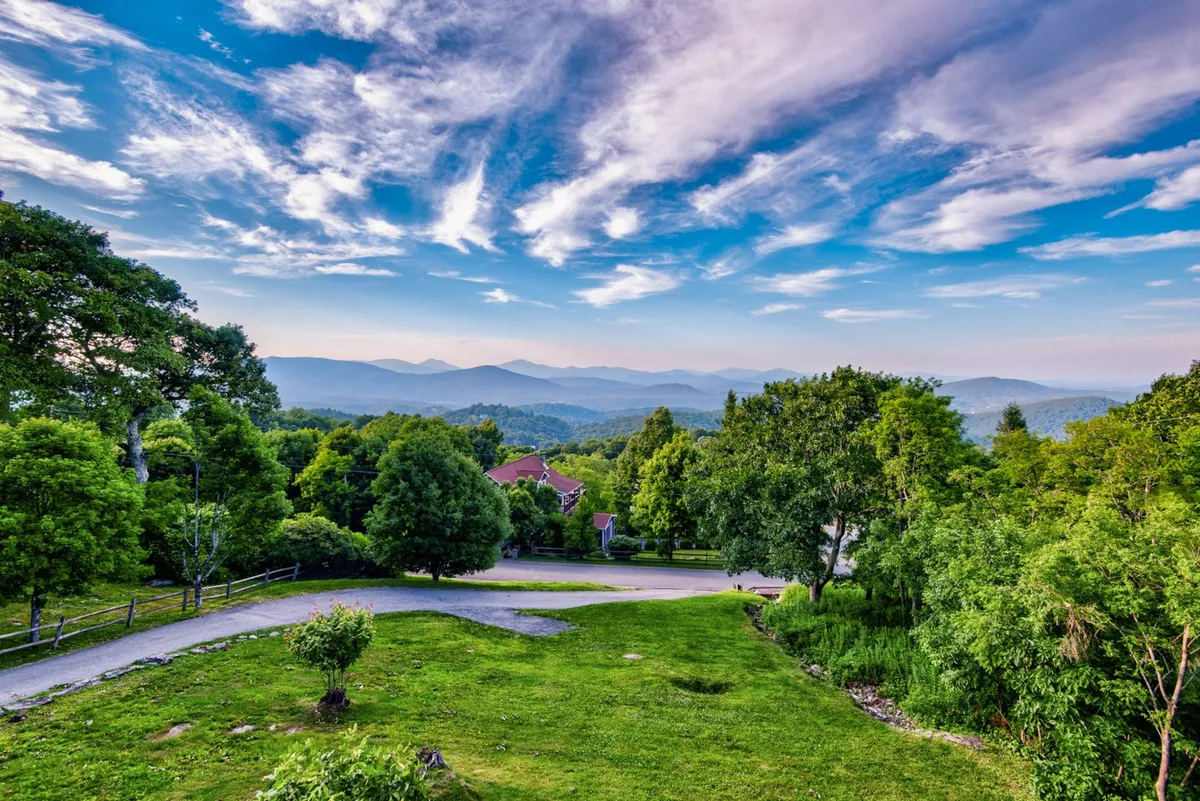 A sweeping view of the mountain range is captured, with lush greenery and a winding road hinting at nearby trails and outdoor adventures. The wide open sky and tranquil atmosphere evoke a sense of exploration and connection to nature.
