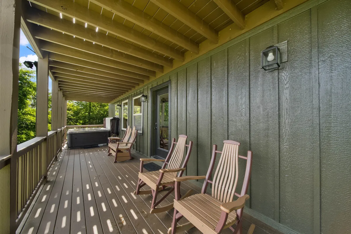 A lower deck scene featuring wooden rocking chairs and a bubbling hot tub, all surrounded by lush greenery. The deck appears serene and secluded, emphasizing the peacefulness and privacy of the outdoor living space. The combination of seating and hot tub aligns with a sanctuary for relaxation in any season.