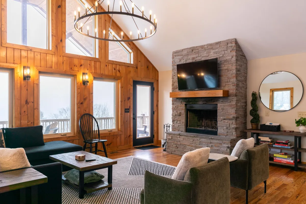 The scene centers on a stone fireplace flanked by large windows that showcase mountain vistas. Comfortable seating creates a cozy atmosphere, enhanced by the warmth of the fire and the inviting glow from the chandelier overhead.