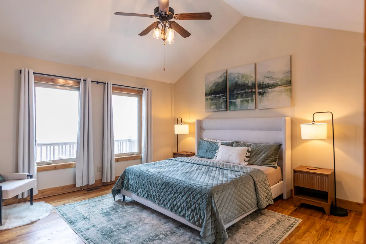 A tranquil King bedroom featuring a large, plush bed with crisp linens, artwork above the headboard, windows allowing natural light, and a side table. The space exudes a sense of peacefulness, inviting restful sleep and quiet mornings.