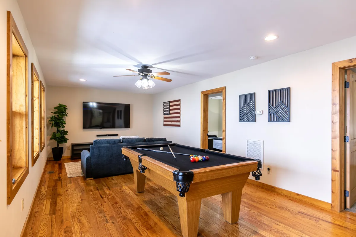 A bright and playful game room featuring a pool table at the center, a comfortable sofa, a large flat-screen television on the wall, and windows bringing in natural light. Wall art adds a creative touch to the relaxed atmosphere.