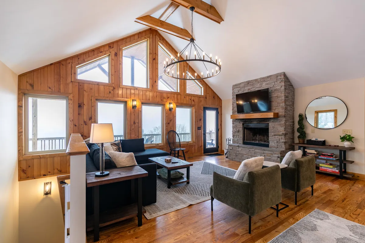 A modern interior living space with large windows framing the mountain scenery, a striking stone fireplace, comfortable seating, and a chandelier overhead. The atmosphere is cozy, ideal for intimate gatherings or quiet moments by the fire.