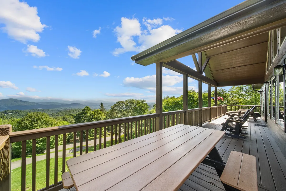 A wide wooden deck with comfortable seating overlooks a sweeping mountain range. The outdoor seating area invites guests to relax or dine alfresco while taking in expansive views. The sky is clear, and the distant mountains are lush and green, evoking a sense of year-round beauty and adventure.