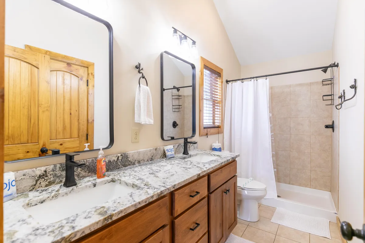 A bright, spa-inspired bathroom with double granite vanities, expansive mirrors, elegant wooden cabinetry, and a spacious shower area. The layout is contemporary and inviting, ideal for relaxing morning or evening routines.