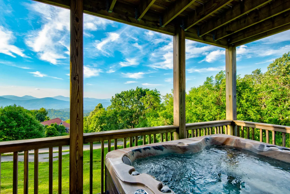 A serene exterior scene featuring a private wooden deck with a four-person hot tub overlooking a breathtaking mountain view. The setting is tranquil and inviting, designed for relaxation and socializing with friends while surrounded by nature.