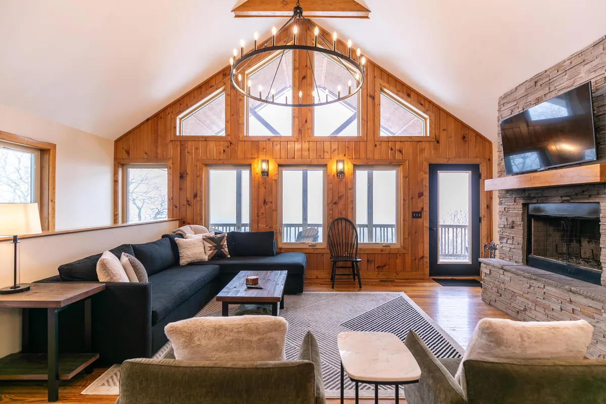 A wide view of the great room captures a plush seating arrangement, dramatic stone fireplace, and tall windows framing the outdoors. Warm wood accents and a statement chandelier add to the inviting, upscale mountain-lodge atmosphere.